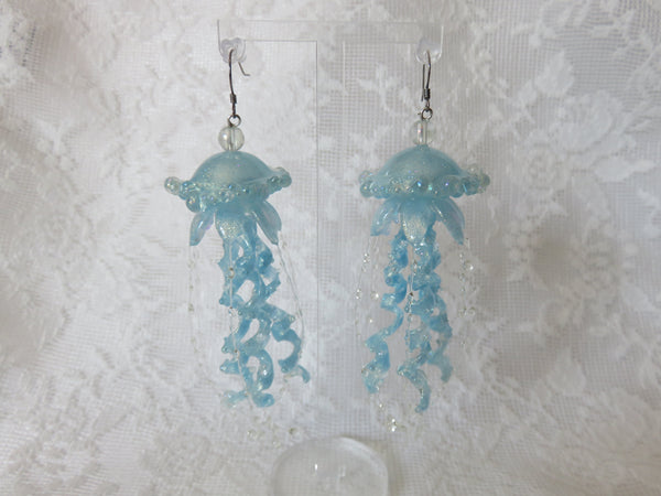 Light blue jellyfish dangle earrings with curled tentacles and small clear bead accents, displayed against a white lace fabric background.