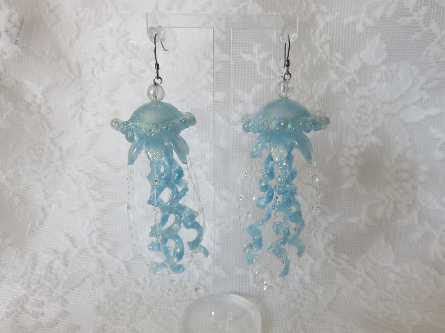 Light blue jellyfish dangle earrings with curled tentacles and small clear bead accents, displayed against a white lace fabric background.